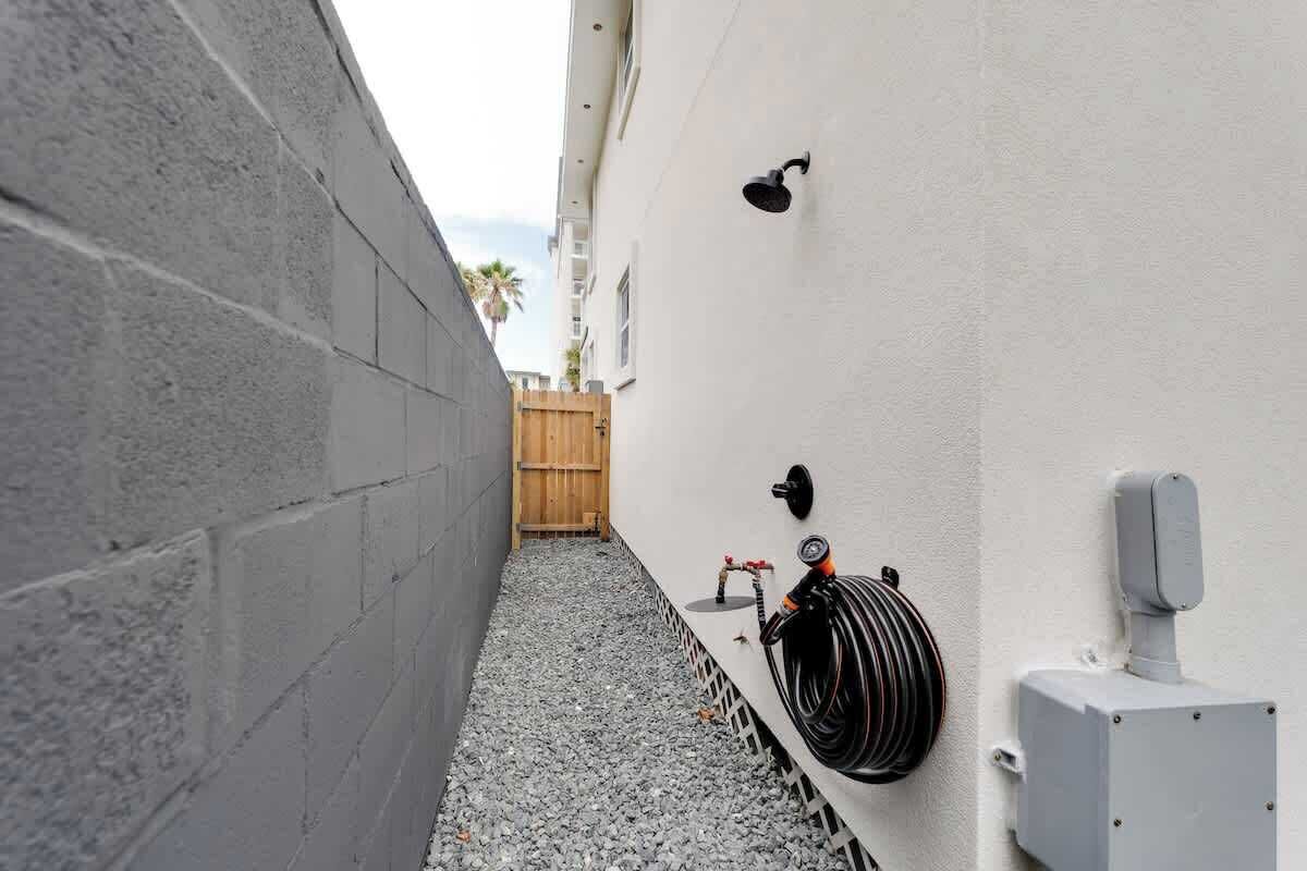 A narrow outdoor corridor with a concrete wall on one side and a white wall on the other, equipped with a hose and a water pump.