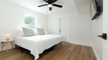 A bedroom with a bed, a ceiling fan, a window, a door, and a wall-mounted TV.