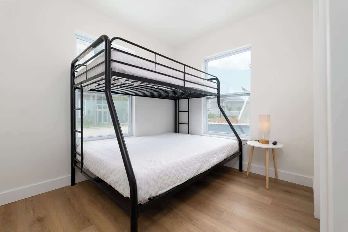 Una cama litera en una habitación con suelo de madera, una pequeña mesa con lámpara, y una ventana que ofrece vistas al exterior.