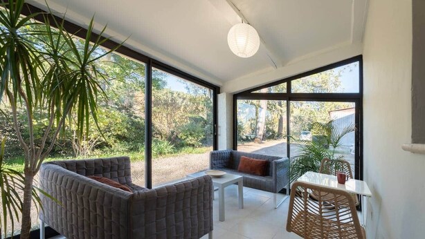 Una habitación amplia con un gran ventanal, un sofá grande con cojines grises, un sofá más pequeño también gris, una mesa blanca y una silla de mimbre.