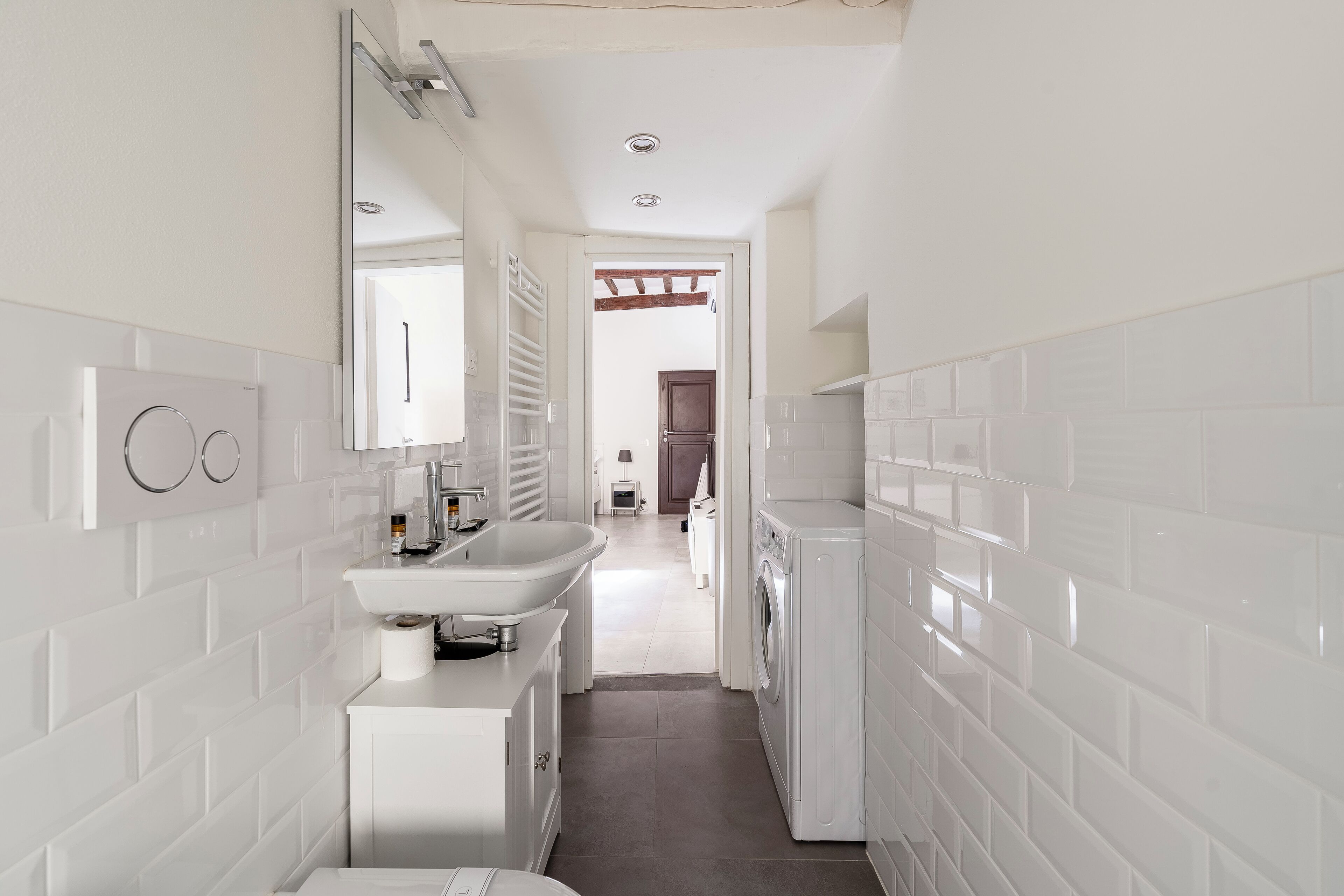 A modern bathroom with white tiles, a sink, a mirror, and a washing machine.
