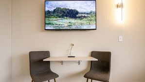 Una habitación con una mesa blanca, dos sillas grises, un cuadro enmarco de un paisaje y una lámpara fijada a la pared.