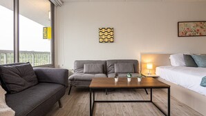 Una habitación de hotel moderna con un sofá gris, una mesa de centro de madera, una cama con ropa de cama blanca y una mesita con una lámpara.