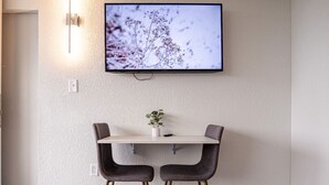 Un comedor moderno con un televisor de pantalla plana en la pared, dos sillas oscuras, una mesita y una planta en maceta.