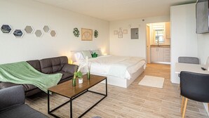 A modern, well-lit room with a sofa, a bed, a dining table, and a kitchen area in the background.