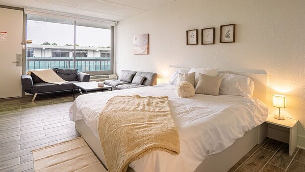 Una camera d'albergo con un letto grande, un divano, un tavolino e un comodino. Presente un balcone con vista sugli edifici.