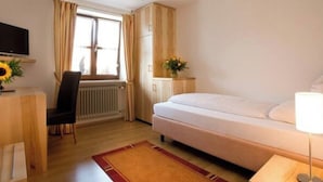 A hotel room with a bed, a desk with a chair, a radiator, a window with curtains, a television, and a vase with flowers. - Boardinghouse calf stretcher (Altdorf)