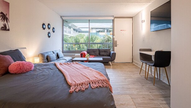 Ein modernes Schlafzimmer mit einem großen Bett, einer Couch, einem Schreibtisch und einem Stuhl. Ein Fenster bietet Blick auf Palmen.