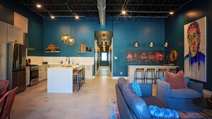 Una cucina moderna con isola centrale, un'area pranzo con sgabelli da bar, un soggiorno con divano e tavolino, e un grande quadro appeso al muro.