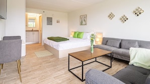 Una habitación moderna y luminosa con una cama, un sofá gris y una mesita. Se ve una zona de cocina al fondo.