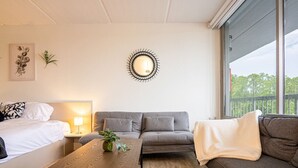 A modern hotel room with a bed, a sofa, a wooden table, and a large window offering a view of the outdoors.
