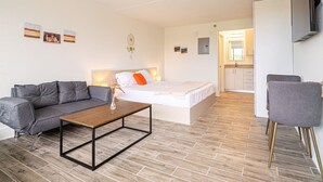 Een moderne hotelkamer met een grijze bank, een houten salontafel, een bed met wit beddengoed en een oranje kussen, een klein tafeltje met een vaas, en een aan de muur bevestigde televisie.
