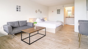 Ein modernes, helles Zimmer mit grauem Sofa, einem Holztisch, einem Bett mit weißer Bettwäsche und einer kleinen Küchenzeile im Hintergrund.