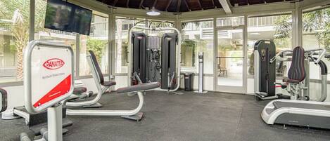 A gym with various exercise equipment, including treadmills and weight machines, under a covered area with large windows.