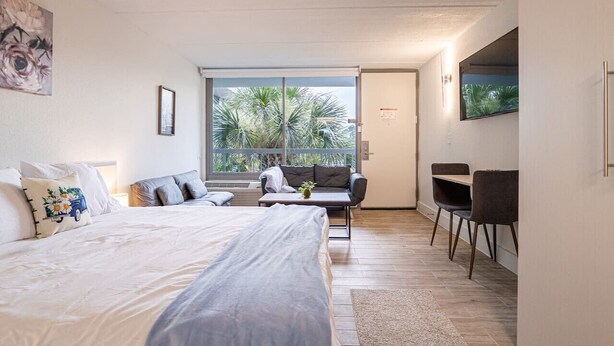 A modern bedroom with a large bed, a sofa, a desk, and a view of palm trees through the window.
