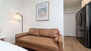 Ein Zimmer mit einem braunen Sofa, einem gerahmten Bild an der Wand, einer Stehlampe und einer Küchenzeile mit einem Kühlschrank.