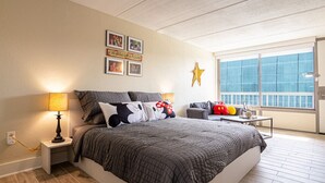 A bedroom with a large bed, a nightstand with a lamp, a small table with toys, and a window with a view of the city. - Kissimmee 1BR Gateway W/pool view  Close to Disney (Kissimmee)