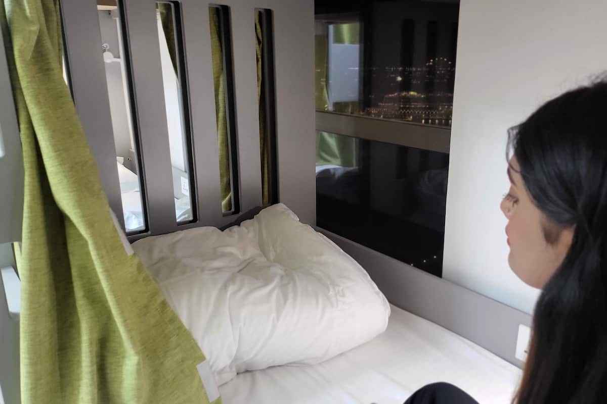 Uma cama com travesseiro branco e cortina verde, uma janela com vista para a cidade e um painel vertical na parede.