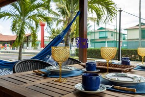 A dining table set with blue and gold dinnerware, a hammock, and a view of palm trees and buildings. - Casa Carneiros 4QTS-JacuzziEChurrasq 039 (Tamandaré)