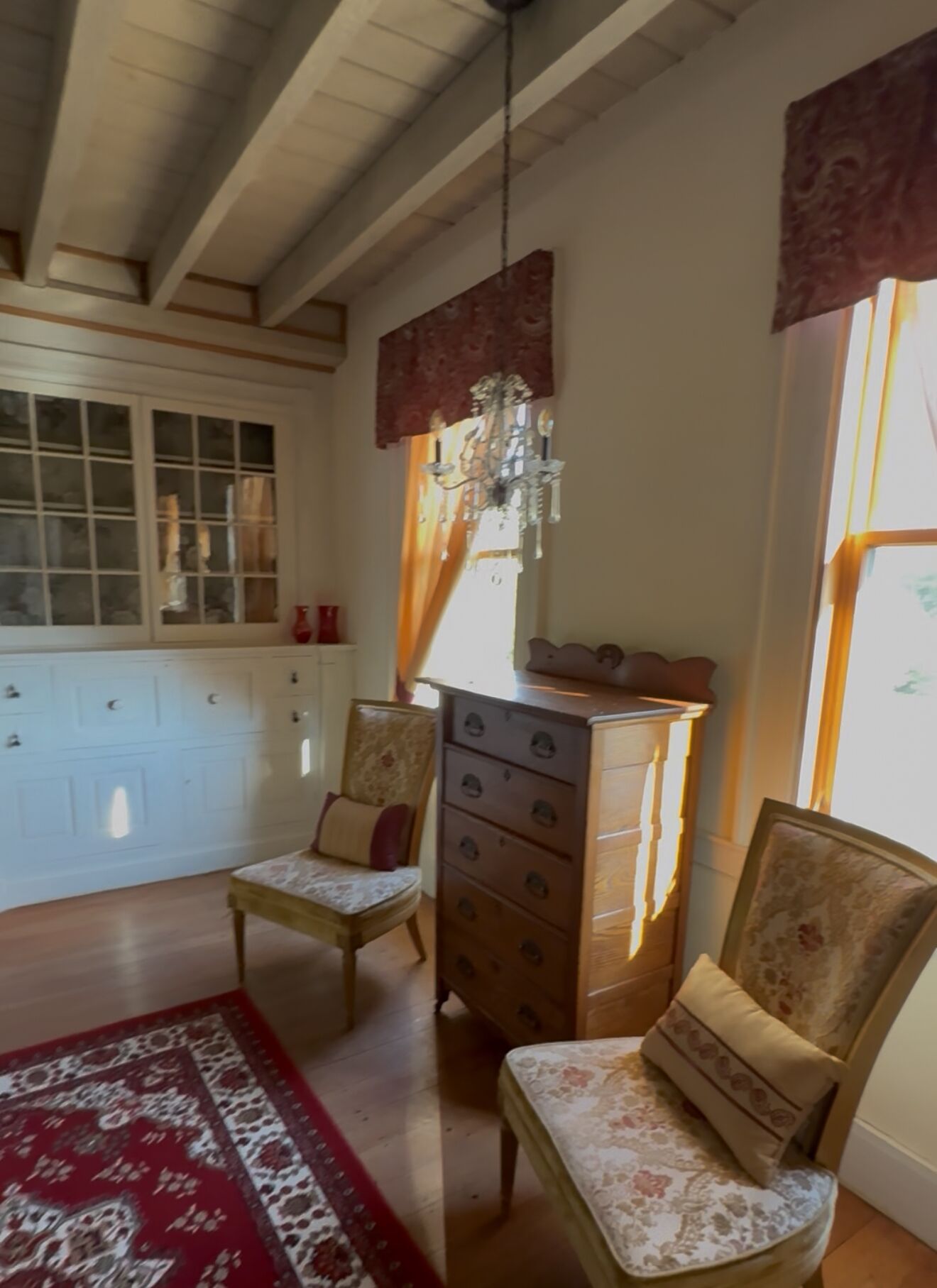 Une pièce avec un plafond en bois, un lustre, une commode en bois, deux fauteuils rembourrés, un tapis rouge à motifs et une fenêtre avec des rideaux.