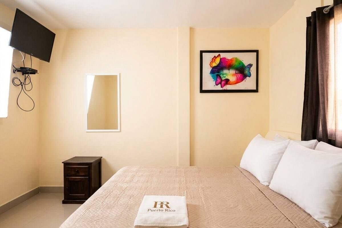 Une chambre d’hôtel avec un lit, un téléviseur fixé au mur, une petite table de chevet, une œuvre d’art encadrée et une fenêtre avec des rideaux.