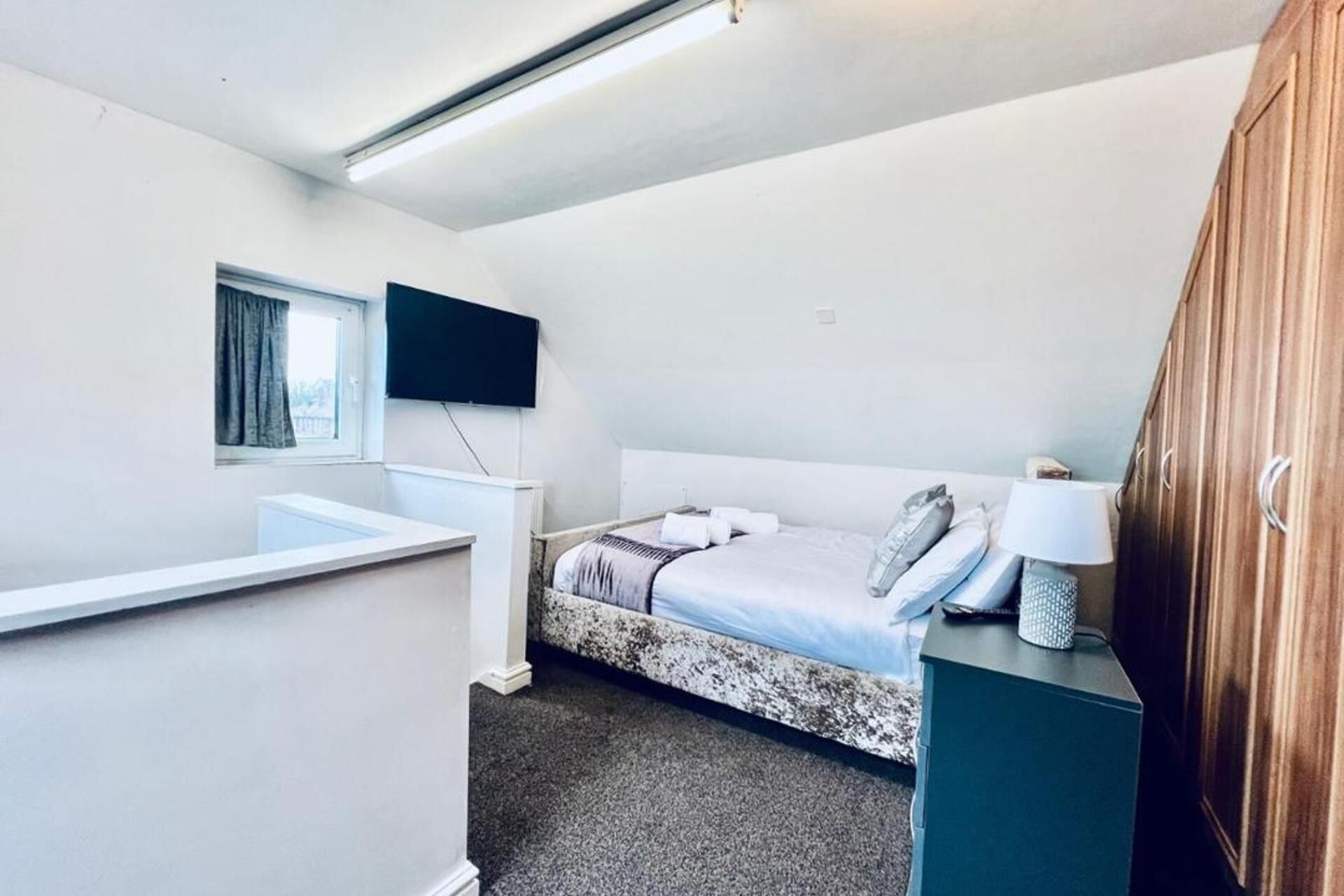 A small, well-lit room with a bed, a TV mounted on the wall, a window with curtains, and a wooden wardrobe.