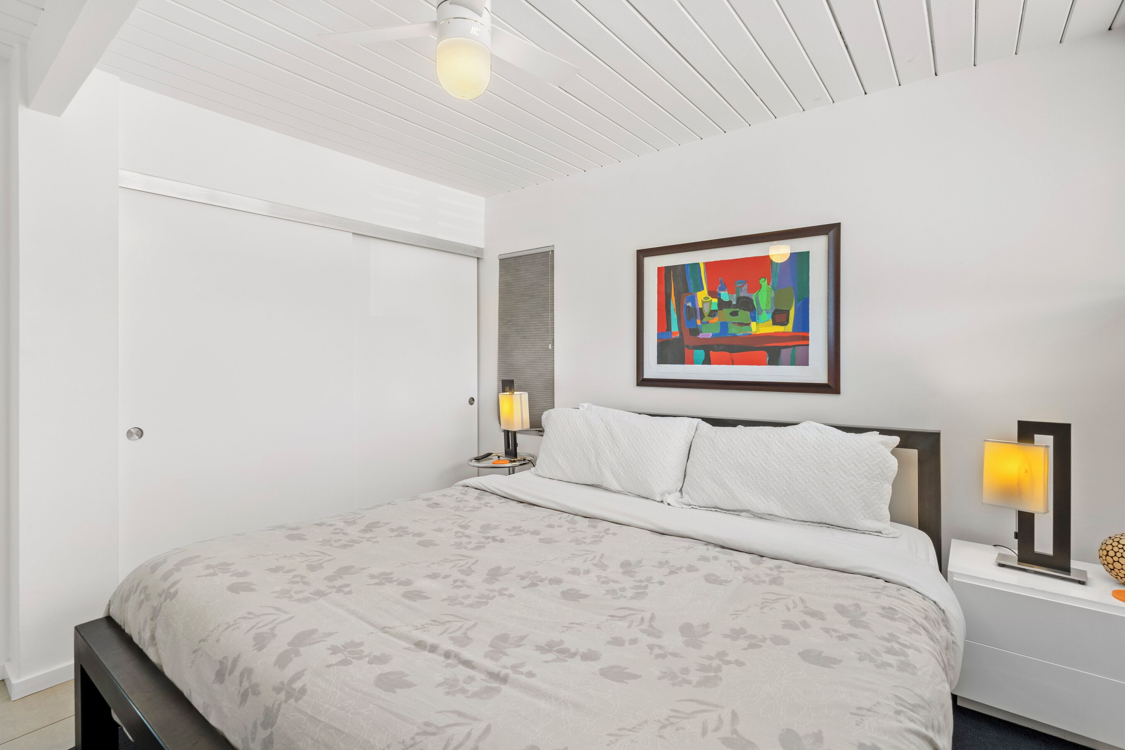 A modern bedroom with a large bed, a nightstand with a lamp, and a colorful abstract painting on the wall.