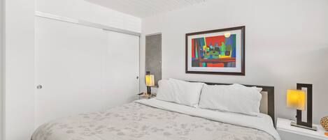 A modern bedroom with a large bed, a nightstand with a lamp, and a colorful abstract painting on the wall.