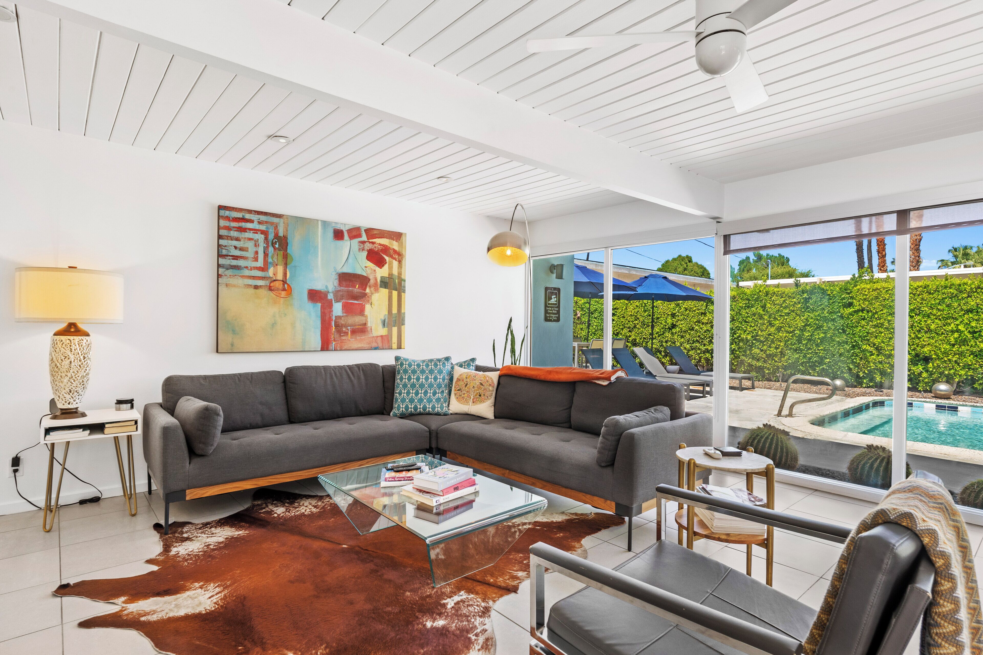 A modern living room with a sectional sofa, glass coffee table, and a cowhide rug. There is a pool visible through large windows.