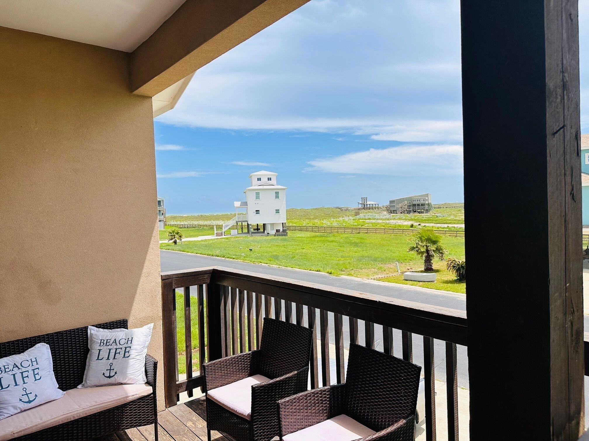 Een balkon met rieten meubels en twee kussens met de tekst 'BEACH LIFE', met uitzicht op een gazon met gebouwen in de verte.