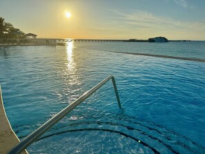 A swimming pool with a sun reflecting on the water, a pier in the background, and a boat docked at the pier.
