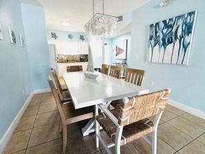 A dining area with a white table, wicker chairs, a chandelier, and a kitchen in the background.