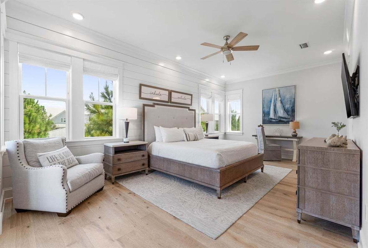 Une chambre avec un grand lit, une table de chevet, une commode et une chaise. On y trouve des fenêtres, un ventilateur de plafond et un tableau au mur.