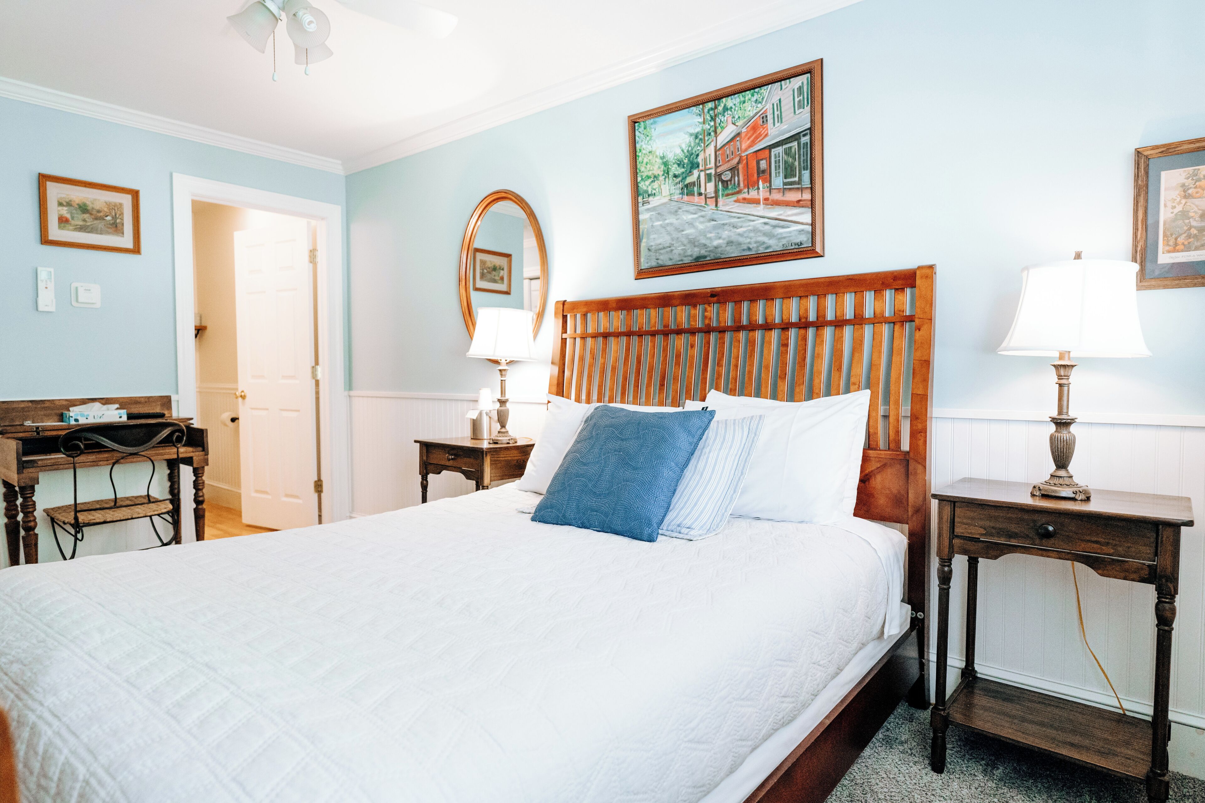 A neatly arranged bedroom with a large bed, two bedside tables, a wooden headboard, a painting on the wall, and a door leading to another room.