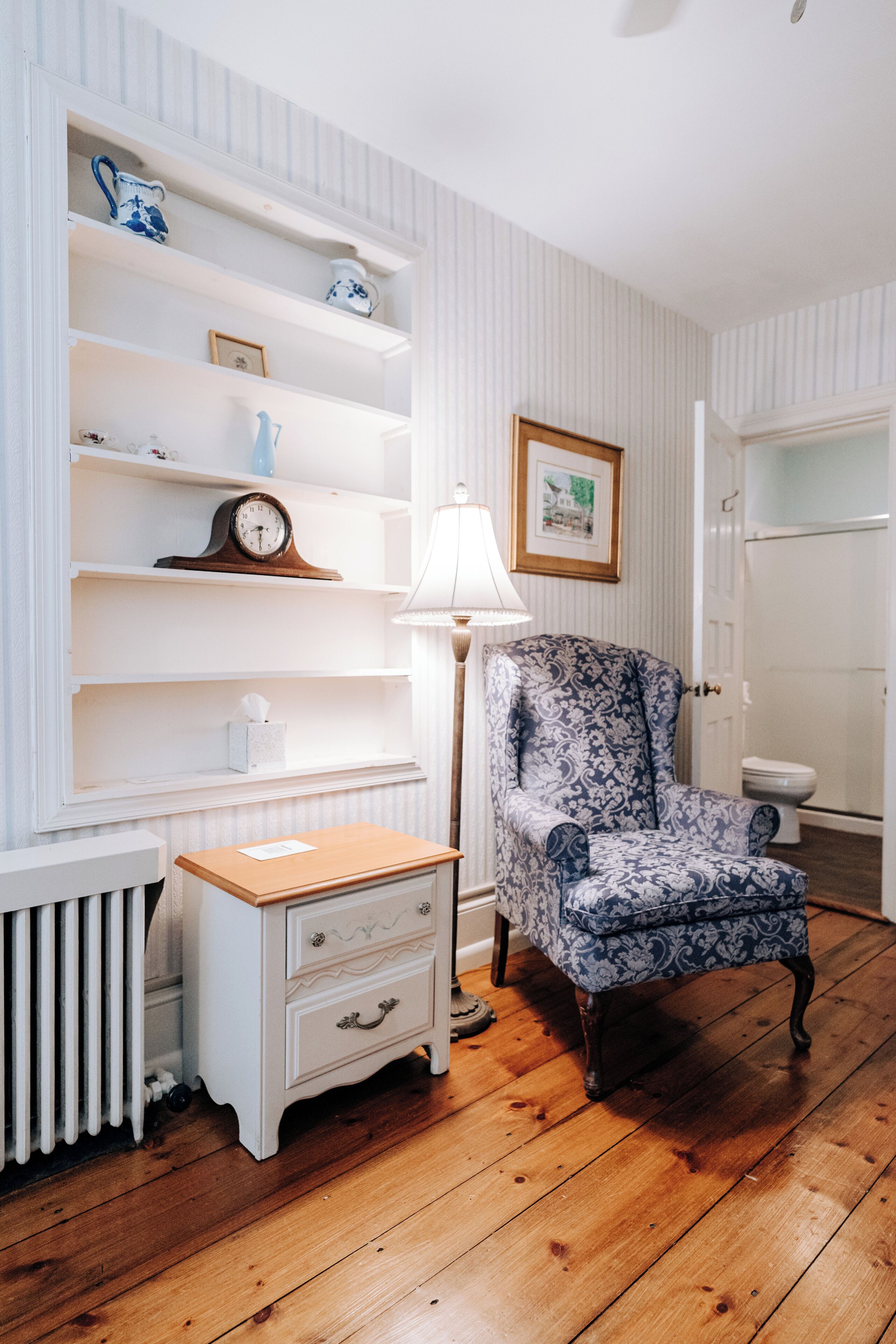 A room with a wooden floor, a white radiator, a white nightstand with drawers, a patterned armchair, a white shelf with decorative items, a framed picture on the wall, and a white door.