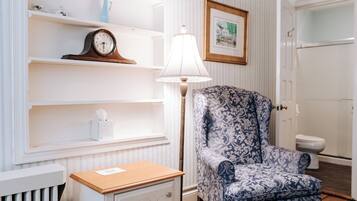 A room with a wooden floor, a white radiator, a white nightstand with drawers, a patterned armchair, a white shelf with decorative items, a framed picture on the wall, and a white door.