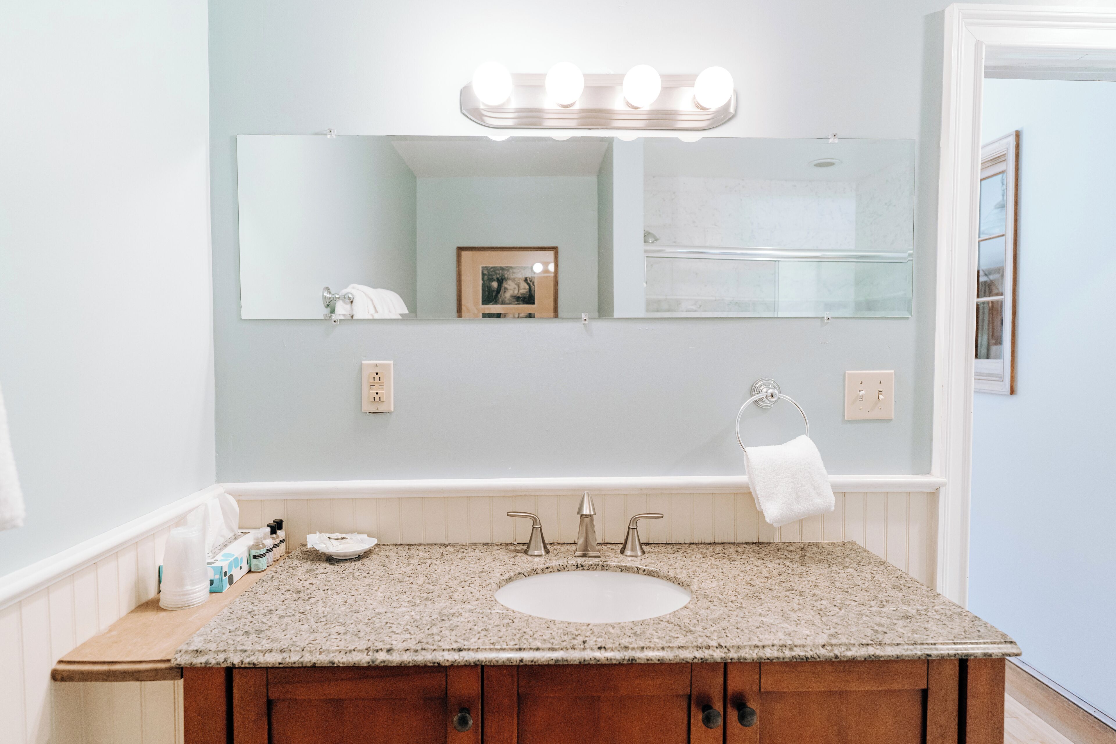 A bathroom with a granite countertop sink, a mirror with lights, a towel rack, and a framed picture on the wall.