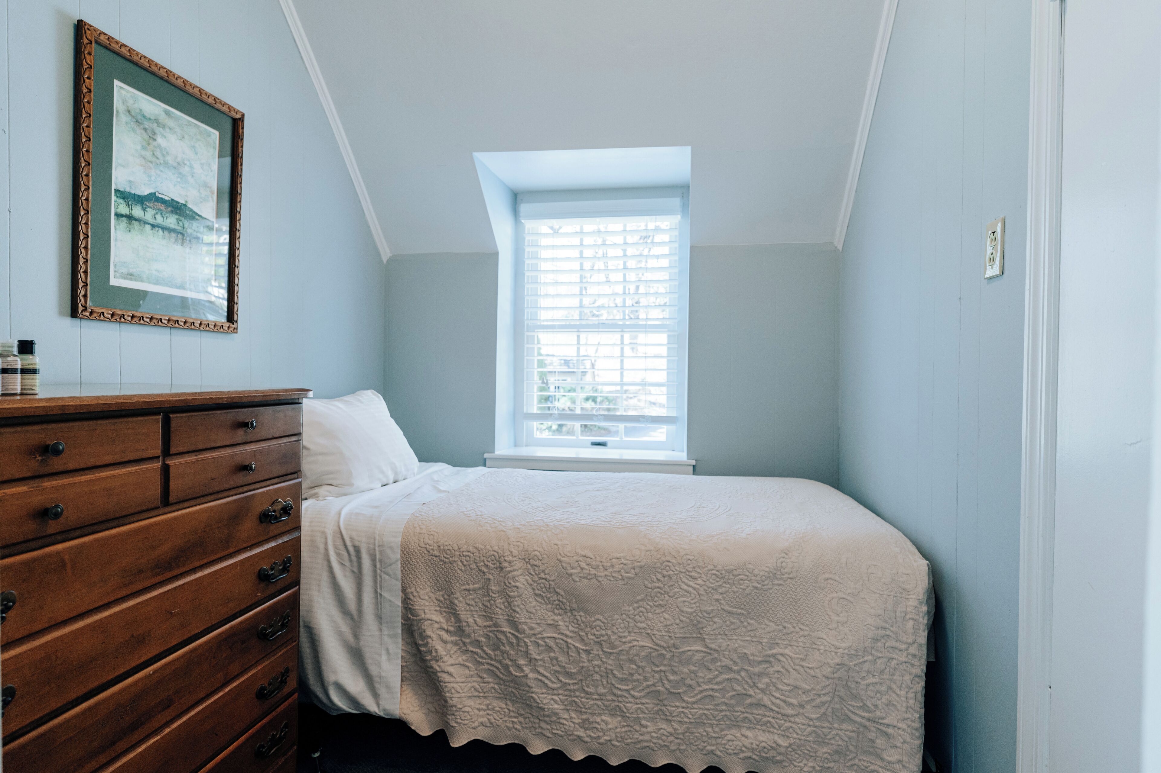 Quarto com cômoda de madeira, cama com colcha branca, janela com persianas e um quadro na parede.