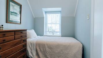 Quarto com cômoda de madeira, cama com colcha branca, janela com persianas e um quadro na parede.