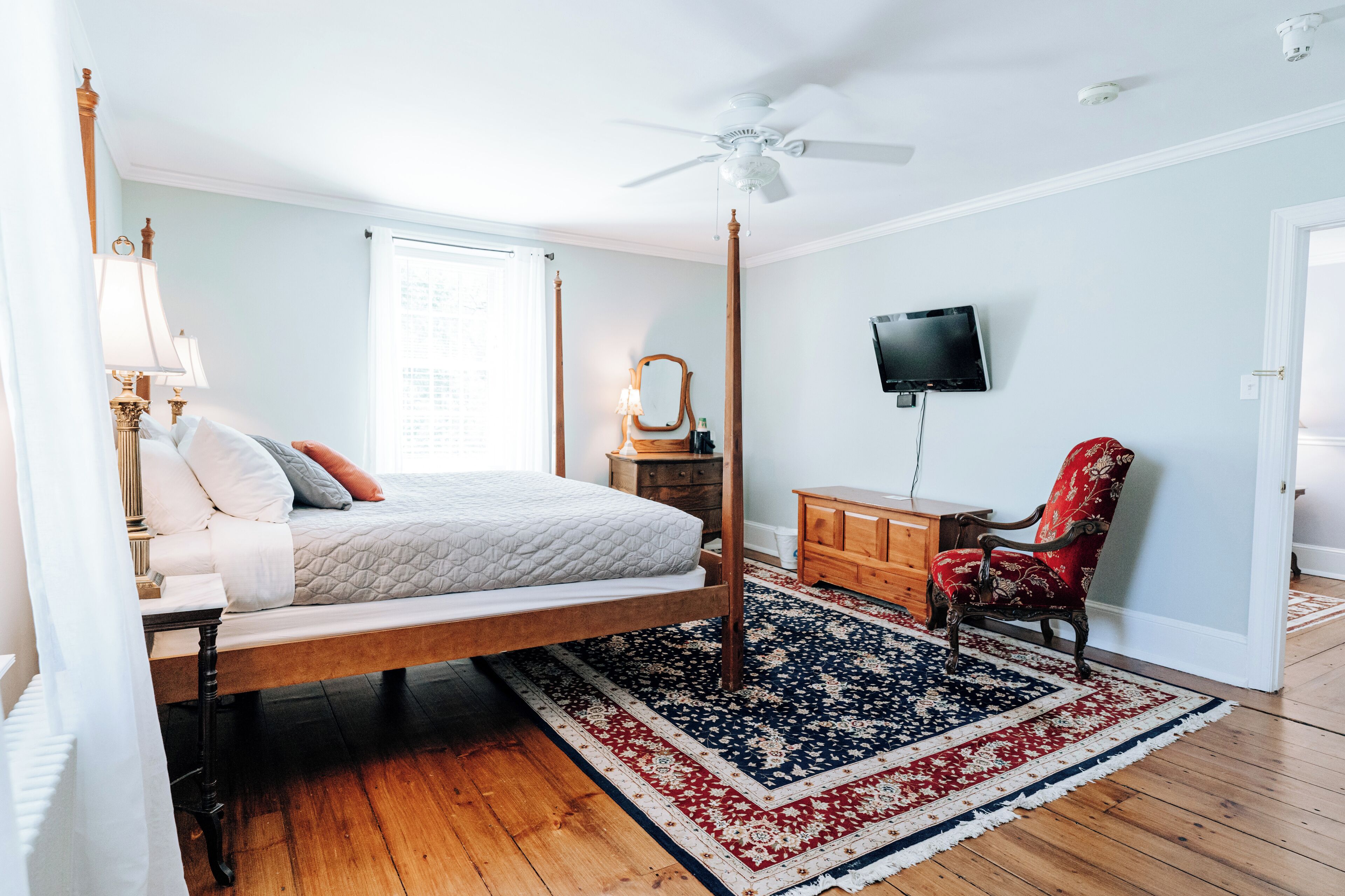 A bedroom with a four-poster bed, a wooden dresser, a red chair, a television on the wall, and a ceiling fan.