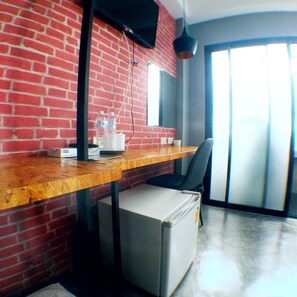 Une pièce avec un mur en briques, un bureau en bois, une chaise, un petit réfrigérateur et une fenêtre avec des stores.