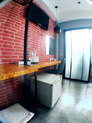 A room with a brick wall, a wooden desk, a chair, a small refrigerator, and a window with blinds.