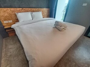 A neatly made bed with white linens and a rolled towel, a wooden headboard, and a view of the outside through a window. - Modern Budget Hotel Hatyai Thailand (Hat Yai)