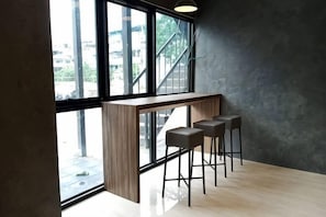 A bar area with a wooden counter and three stools, large windows, and a view of buildings outside. - Modern Budget Hotel (Hat Yai)