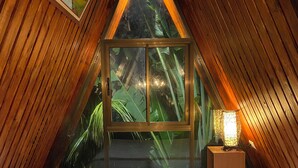 Een gezellige, driehoekige kamer met een bed, een houten plafond en een groot raam met uitzicht op groen.