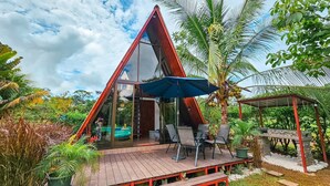 Een driekantig glazen huis met een houten terras, zitplaatsen buiten en een zwembad.