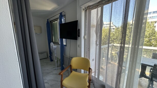 Un balcon avec une chaise jaune, un téléviseur fixé au mur et une vue sur l’extérieur.