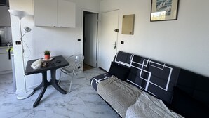 Une petite chambre moderne, équipée d’un lit, d’un bureau et d’une chaise. On trouve sur le bureau une plante en pot, une lampe et, au mur, un tableau encadré.