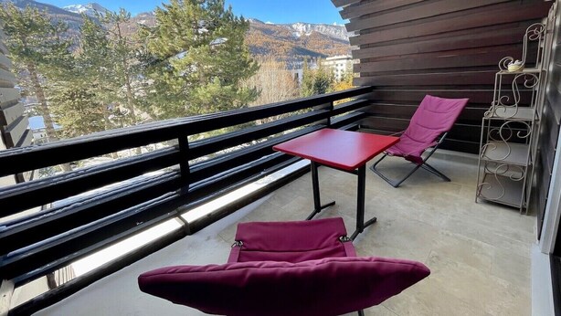 Varanda com mesa vermelha e duas cadeiras, uma estante de metal e vista para montanhas e árvores.