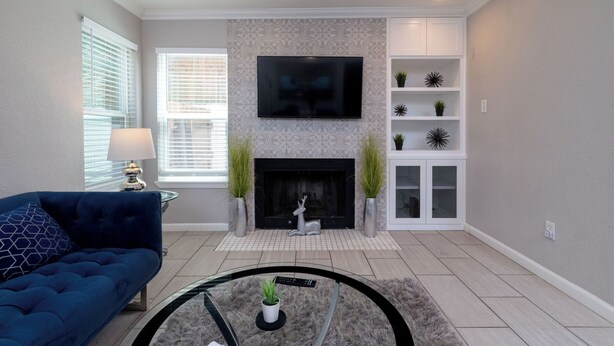A living room with a fireplace, a flat-screen TV, a blue sofa, and a glass coffee table with a remote control. - Patio + Covered Terrace - Steps to TCU (Fort Worth)